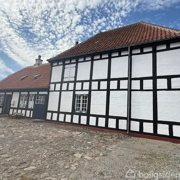 Bindingsværkshus med hvide mure og rødt tegltag, står på en stenbelagt gårdsplads. Himmelen er blå med få skyer. Træer ses i baggrunden. Ingen tekst er til stede.