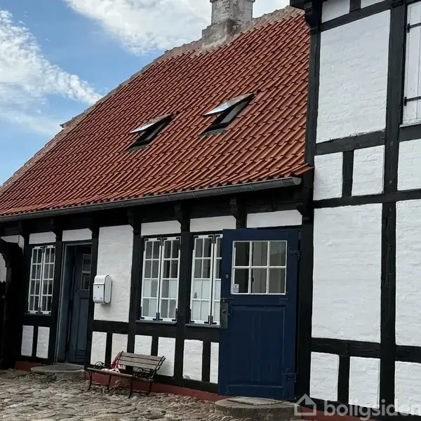 Et bindingsværkshus med hvidmalede vægge og sorte træbjælker. Det har rødt tegltag og små vinduer. Foran huset er en brostensbelagt gårdsplads, der skaber en idyllisk atmosfære.