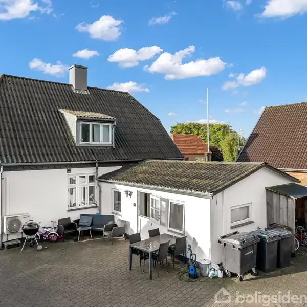 En enkeltseng er uredt i et rum med hvidmalede vægge og trægulv. Ved siden af står et lille bord med ting på. Et vindue med persienner og en airconditionenhed er på væggen. En tv-skærm er placeret på gulvet.