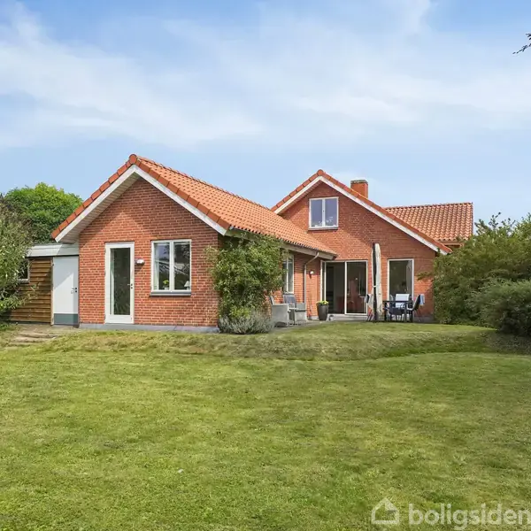 Et rødt murstenshus med et rødt tegltag står på en grøn græsplæne omgivet af buske og træer. Foran huset er der en terrasse med havemøbler.