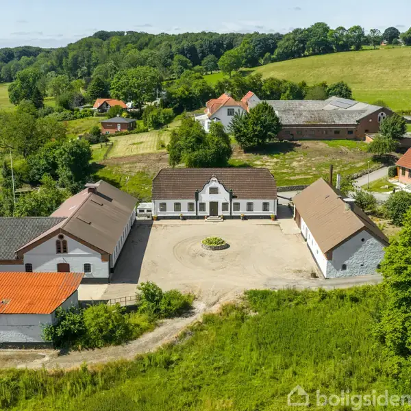 En gård har en central gårdsplads omkranset af hvide bygninger med mørke tage. Den omgivende landskab er grønt med bakker og skove under en klar himmel.
