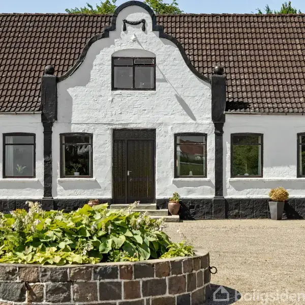En hvid facadebygning med mørkt saddeltag står foran en grusplads. Foran bygningen er en rund stenkantet planteboks fyldt med grønne planter. Ingen tekst er til stede.
