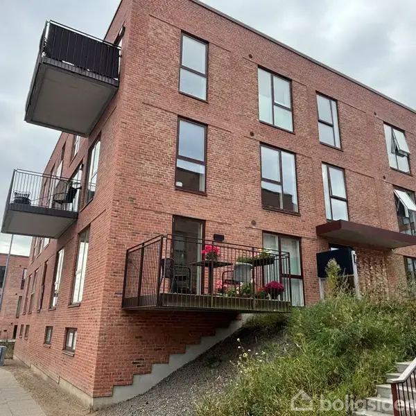 Rødt murstenslejlighedskompleks med flere etager og altaner, nogle steder med blomster og møbler. Til venstre en trappe, der fører op til en altan. Overskyet himmel.
