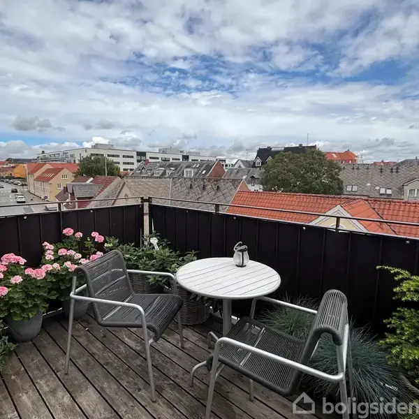 En terrasse med et rundt bord og to stole omkring det; omgivet af blomster i potter. Udsigt over hustage og en by bagved under en delvis skyet himmel.