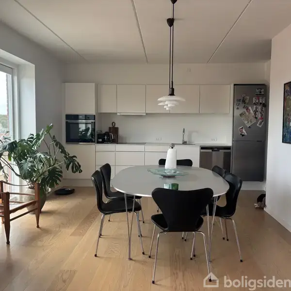 Et lyst køkken med moderne indretning har et rundt spisebord med seks sorte stole. Køkkenet har hvide skabe, en ovn og et stål køleskab. En stor plante står ved vinduet.