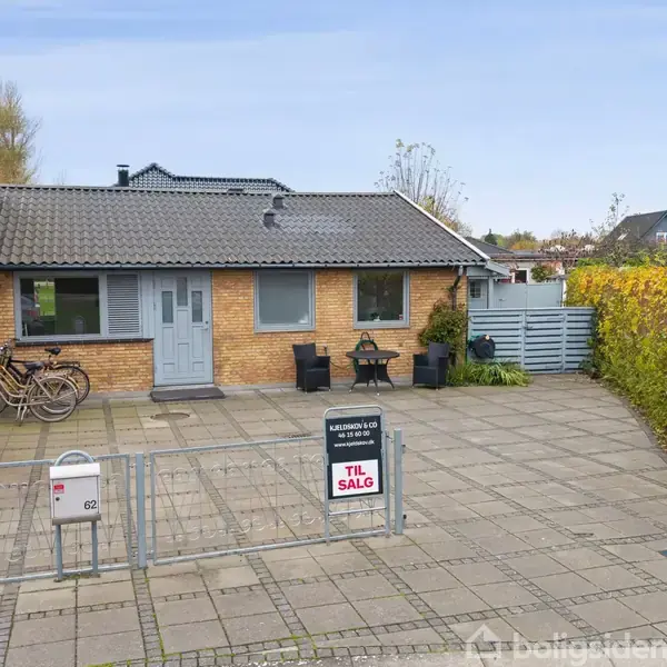 Et murstenshus står bag et hegn, med en cykel og bord ved siden af. Et "Til salg" skilt står i indkørslen. Fliser dækker gårdspladsen, omgivet af buske.