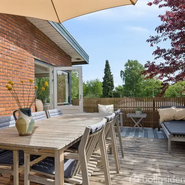 Terrasse med bord og stole under en parasol. En sofa med puder står ved siden af på trægulvet. Murstensvæg til venstre, haveomgivelser med træer i baggrunden.