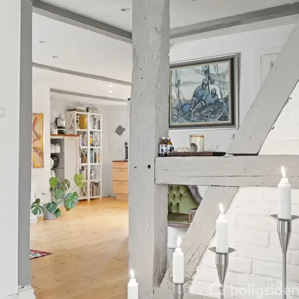 Et interiør med en hvidmalet bjælke og stearinlys i forgrunden. Flere farverige malerier og planter pryder det lyse træinteriør med åbne hylder i baggrunden.