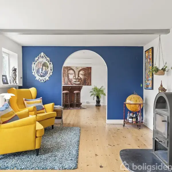 To gule lænestole står stille på et blåt tæppe i en lys stue, hvor en brændeovn er placeret ved siden af en blå væg med en bueåbning til et andet rum.