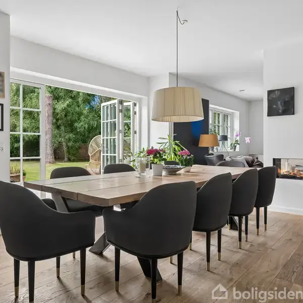 Spisebord med sorte stole står i en lys, moderne stue. Dobbelt franske døre åbner ud til en grøn have. Væggene er pyntet med billeder, og der er en pejs.