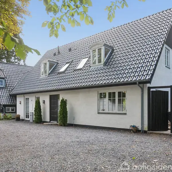 Hvidt hus med sort tagstensbelægning, ingen synlig handling, beliggende i en grusbelagt gårdsplads ved siden af træer og buske. En bil er parkeret til venstre.