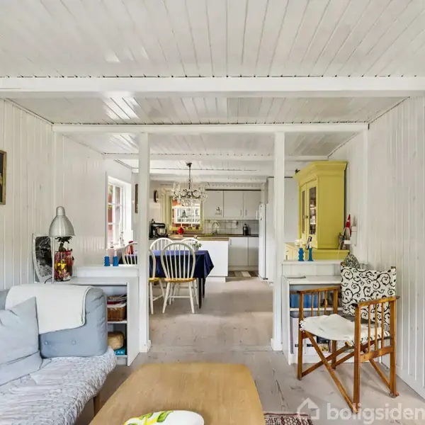 Stue med sofa og træborde, åbner op til spisestue med stole og blåt dækket bord. Lys malet trævægge og loft, skaber en lys, hyggelig atmosfære.