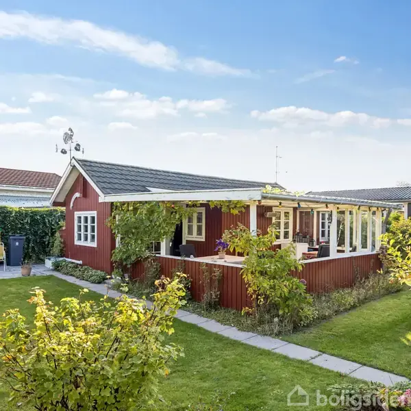 Et rødt hus med hvide vinduer på en solrig dag. Huset har en overdækket terrasse med planter. Det står i en velplejet have med græsplæne og buske.