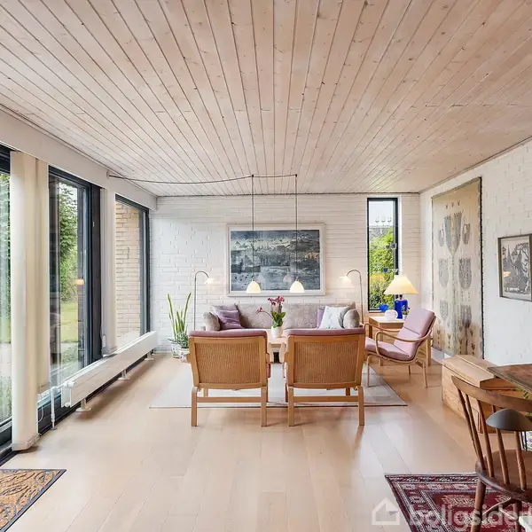 Stuen har en sofagruppe med lyse træstole og bord. Rummet er lyst med store vinduer og hvide vægge. Der er lamper, et skrivebord, og dekorerende billeder på væggene.