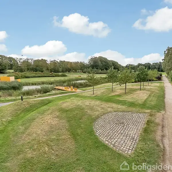Græsplæne strækker sig langs en grussti, omgivet af træer og buske i en park. En træbro krydser over en lille dam. Himlen er blå med spredte hvide skyer.