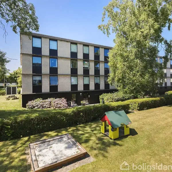 Lejlighedsbygning med ensartede vinduer står stille i grøn, solrig park med træer. I forgrunden ses en lille legeplads med sandkasse og farverigt legehus.