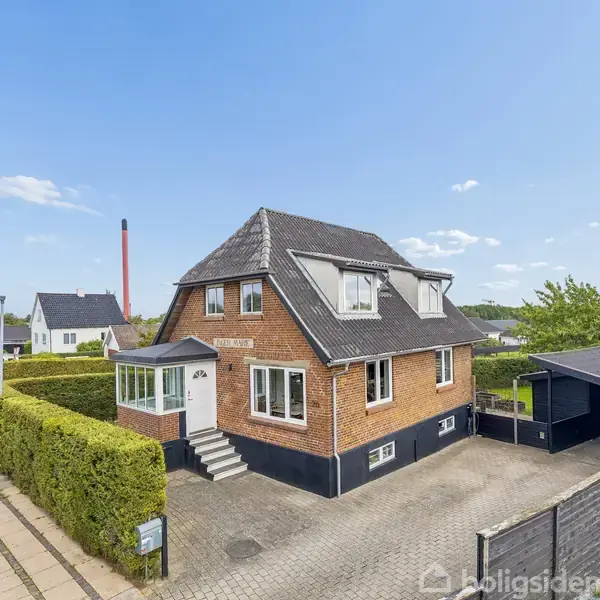 Et rødt murstenshus med en trappe til hoveddøren, omgivet af velplejede hække og en flisebelagt indkørsel, ligger i et villakvarter med blå himmel og spredte skyer.