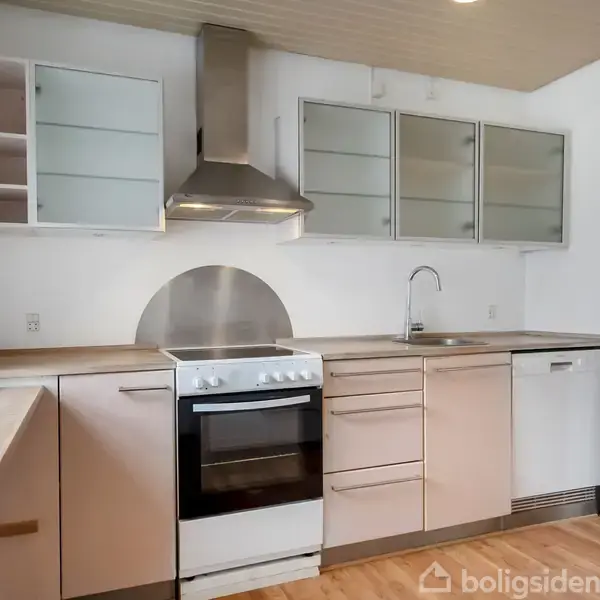 Et køkken med hvide skabe, en komfur med rustfri stål emhætte, vask, og opvaskemaskine. Det har trælaminatgulv, lys vægmaling, og åbner op til en stue gennem en bueformet indgang.