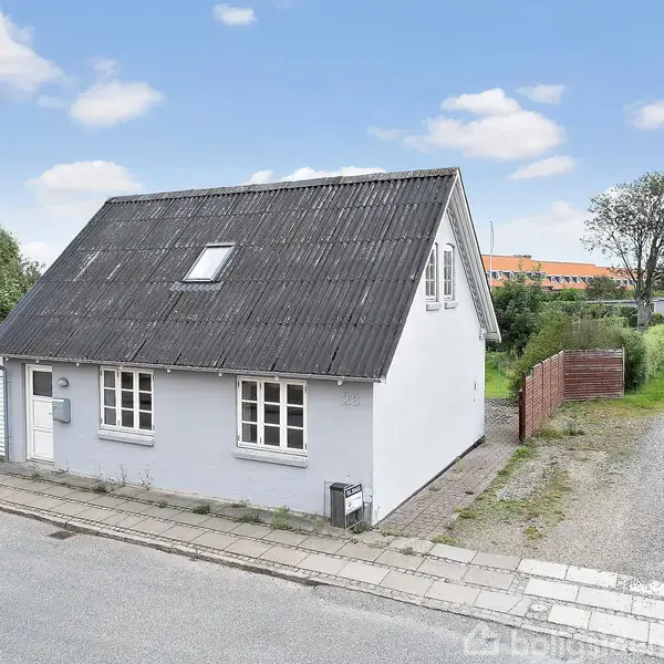 Et lille hvidt hus med sort tag står ved en vej, omgivet af en grussti og buske. Huset har flere vinduer og et ovenlysvindue under en klar, blå himmel.