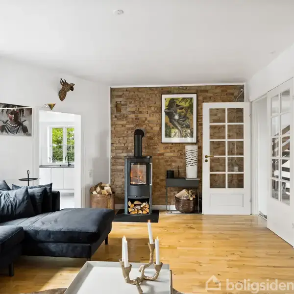 En mørk sofa står i en stue mod en murstensvæg med en brændeovn. Ved siden af er der glasdøre, kunst på væggene, og et træbord med lysestager.