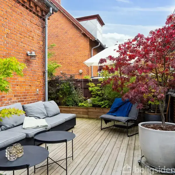 Grå sofa står på en træterrasse, flankeret af små sorte borde. Omkring er rødbrune murstensvægge, grønne planter og et stort, rødt løntræ i krukke.