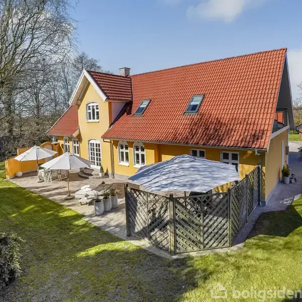 Et gulmalet hus med rødt tegltag står i en have med græs og træer. Et terrasseområde har parasoller og havemøbler. Sollys skinner gennem de omkringliggende træer.