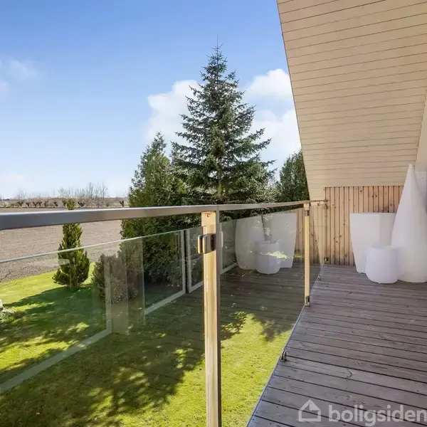 Balkon med glasrækværk strækker sig langs en bygning; omgivet af grøn græsplæne og træer, står to hvide dekorative krukker i hjørnet nær glasskydedøre.