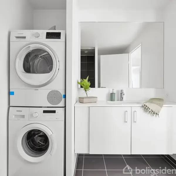 Vaskemaskine og tørretumbler står stablet til venstre. Badeværelsesskab med spejl til højre, omgivet af hvide vægge og sort gulv. Håndklæde hænger på væggen.