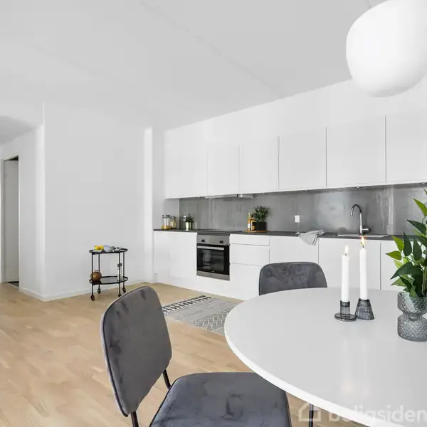 Spisebord dækket med to lysestager og en plante står i en moderne, rummelig stue-køkken kombination. Hvide elementer og åben indgang til anden del af lejligheden.