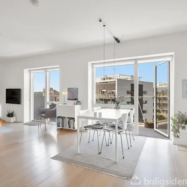 Et lyst, moderne rum med et hvidt spisebord og stole stående på et tæppe. Døre åbner til en balkon, omgivet af store vinduer med udsigt over andre bygninger. Plante i hjørnet.
