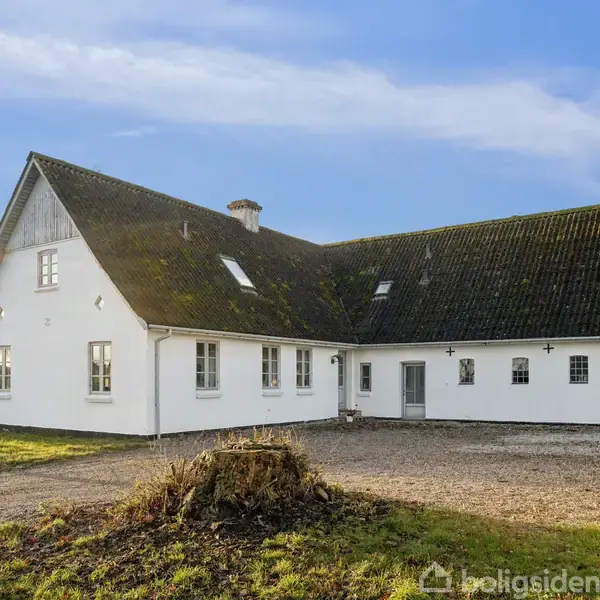 Et hvidt, vinkelformet hus med mørkt tag står på en gårdsplads omgivet af græsplæner under en blå himmel.
