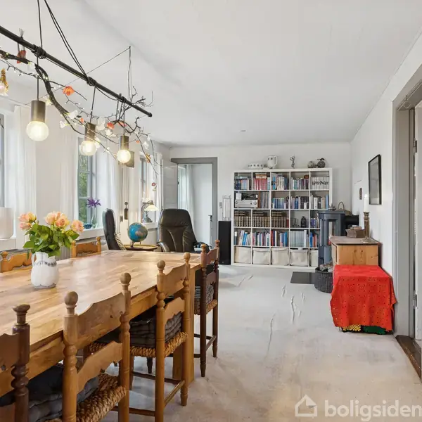 Langt spisebord med træstole står under en lysinstallation med pærer. Bordet er dekoreret med blomster i vase. Rummet har hylder fyldt med bøger og lyse vægfarver.