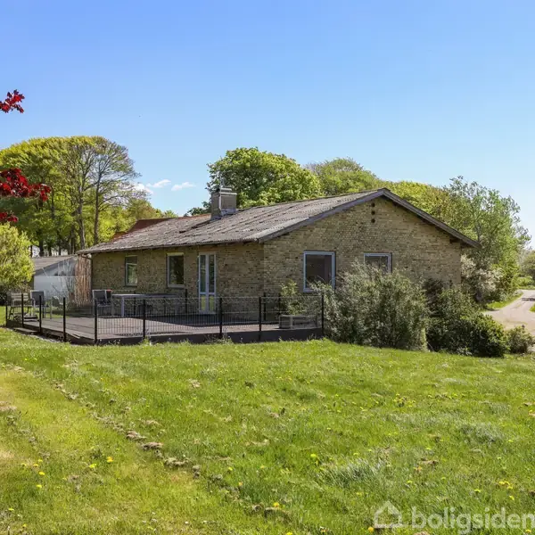 En stenhus med terrasse står i en grøn have omkranset af træer. En grusvej fører til og fra huset. Det er en solrig dag med blå himmel.