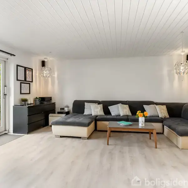 Stor grå hjørnesofa med puder står i en lys stue, sofabord foran. Store vinduer til venstre viser have. To lamper hænger fra loftet, indrammet kunst til højre.