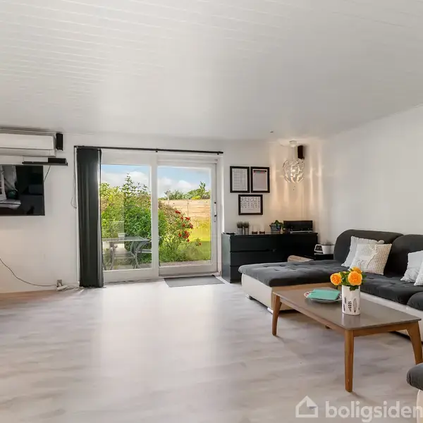 Sofa med puder står i en stue ved siden af et sofabord med blomster. Glasdøre leder ud til en have. Spisebord med stole og et fjernsyn ses i baggrunden.