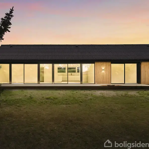 Et moderne hus med store glaspartier lyser op i skumringen i en have omgivet af træer. Indendørs belysning bidrager til den varme atmosfære.