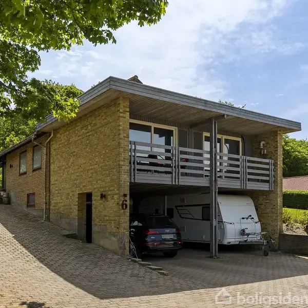 En murstensvilla med integreret carport, hvor en bil og campingvogn holder parkeret. Omkring huset er der grøn beplantning. På facaden ses tallet "6".
