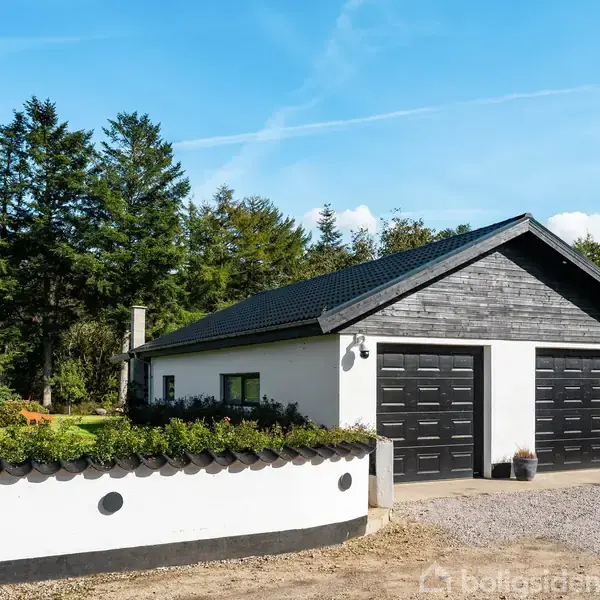 En hvid garagebygning med sort tag og to sorte porte står stille omgivet af træer. Foran er der en grusbelagt indkørsel og en hvid mur med planter.