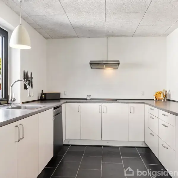 Et moderne køkken med hvide skabe og sorte bordplader. To pendellamper hænger over hjørnet. Sorte fliser dækker gulvet. Et vindue til venstre bringer naturligt lys ind.