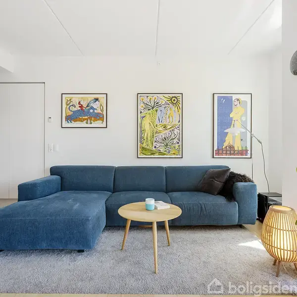 En blå sofa står centreret på et gråt tæppe. Et rundt træbord står foran sofaen. Væggene har tre farverige malerier, og en plante står ved siden af en lampe i hjørnet.