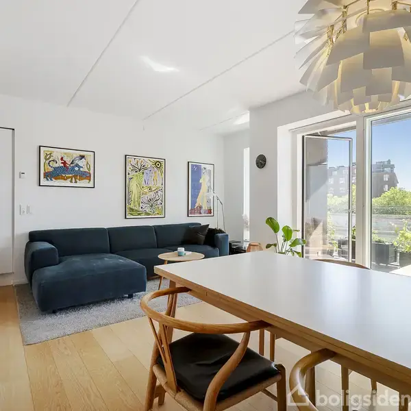 En moderne stue med en mørkeblå sofa, der står på tæppe. Bord med stole står foran åben altandør med udsigt til grønne planter. Væggene har farverige billeder.