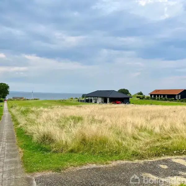 En moderne sort hus står i en åben mark. Græsset er langt og vejret er overskyet. Et stisystem fører mod havet i horisonten, omgivet af grønne træer.