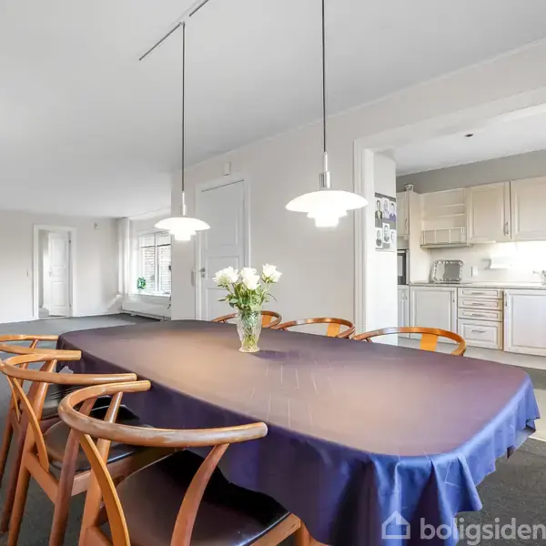 Et spisebord med blå dug og træstole er omkranset af lamper. En vase med blomster står på bordet. I baggrunden ses et køkken med hvide skabe og en åben dør.