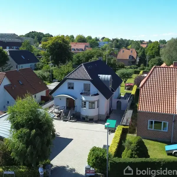 Et hvidt hus med en sort tagbelægning står i en gårdsplads med brosten og parkeringsplads. Et tilstødende skur til venstre har parkerede biler og en bænk.