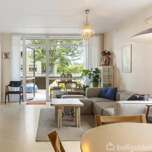 Stuen indeholder en beige sofa med puder, træbord foran, og åbner ud til en terrasse med bord og stole. Store vinduer giver udsigt til en frodig have.