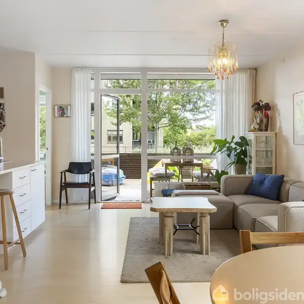 Stue med sofa og sofabord, åbner ud mod en terrasse gennem store glasdøre. Køkkenhjørne med barstole til venstre, spisebord i forgrunden, og grønne planter omkring.