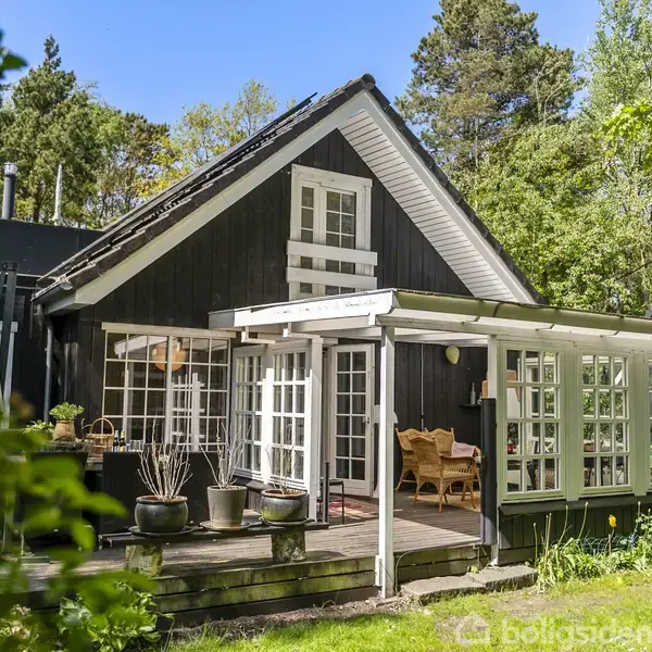 Sort port stående åben; stenvej fører mod sort hus med terrasse og hvide møbler; omgivet af træer og buske. Husets nummer "7" er synligt på højre side.