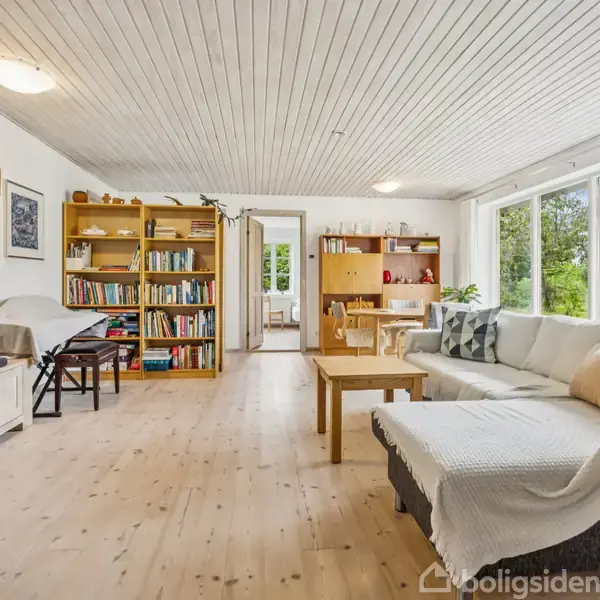 Stue med en stor, grå sofa ved et træbord, placeret på et lyst trægulv. Reoler med bøger langs væggen, og store vinduer, der viser en grøn natur udenfor.