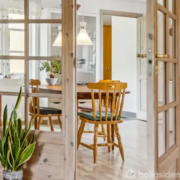 En åben trædør med glasruder viser et lyst køkkenmiljø. Et rundt spisebord med træstole står centralt, omgivet af planter og lamper. En slank potteplante står tæt på døren.