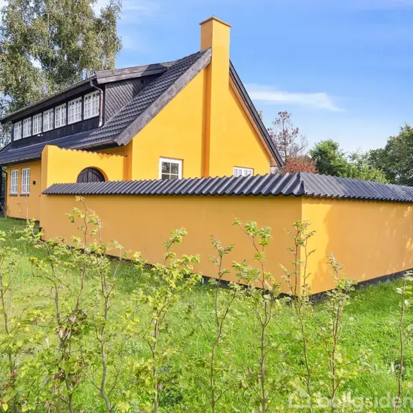 Gult hus med sort tag står i en frodig have omkranset af grønne buske og træer. Klar blå himmel ses i baggrunden, hvilket skaber en fredelig atmosfære.